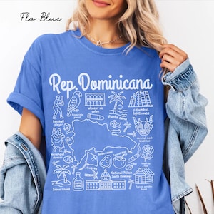 May include: A blue short-sleeved t-shirt featuring a white graphic map of the Dominican Republic surrounded by illustrations of a baseball player, parrot, carnival mask, palm trees, and landmarks with text labels including Rep. Dominicana.