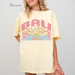 May include: A pale yellow short-sleeve t-shirt featuring a distressed graphic print with the text "BALI" and "Indonesia". The design includes a stylised sun, palm trees, and ocean waves in shades of pink, green, and blue. The word "Banana" is printed in the top left corner.