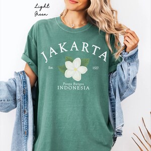Op de afbeelding: Een lichtgroen t-shirt met de witte tekst JAKARTA, Est. 1527 en Puspa Bangsa INDONESIA. Een centrale afbeelding toont een witte bloem met groene bladeren. Het t-shirt wordt gedragen met een blauw spijkerjack en de tekst Light Green is zichtbaar.
