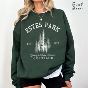 May include: A forest green crewneck sweatshirt featuring white screen-printed text that reads ESTES PARK, EST. 1859, Gateway to Rocky Mountain, COLORADO, and a graphic illustration of a cluster of pine trees centered on the chest.