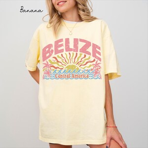 May include: A pale yellow short-sleeve t-shirt featuring a graphic print with the text BELIZE and Central America. The design includes a stylized sun, palm trees, and ocean waves in shades of pink, green, and blue with a distressed texture.