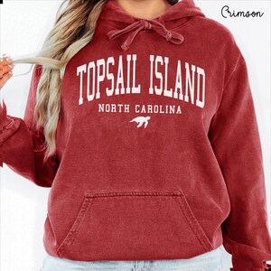 May include: Crimson red hoodie with a drawstring hood and a front pocket. The hoodie features the text "TOPSAIL ISLAND NORTH CAROLINA" in white block letters, with a white turtle graphic below the text.