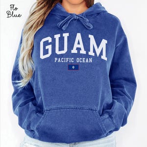 May include: A blue hooded sweatshirt featuring the word "GUAM" in large white letters. Beneath, it reads "PACIFIC OCEAN" with a small Guam flag. The sweatshirt has a front pocket and a drawstring hood. The colour is described as Flo Blue.