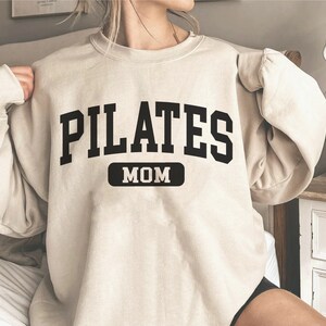 Pu&ograve; includere: Una felpa beige con la scritta "PILATES MOM" in lettere nere. Il testo &egrave; in un font in stile universitario.