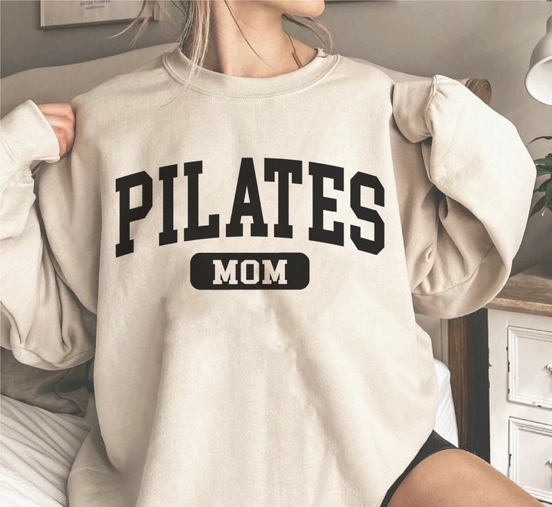 Pu&ograve; includere: Una felpa beige con la scritta "PILATES MOM" in lettere nere. Il testo &egrave; in un font in stile universitario.