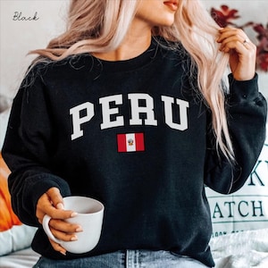 Könnte beinhalten: Ein schwarzes Langarm-Sweatshirt mit Rundhalsausschnitt, das das Wort PERU in großen, weißen Großbuchstaben bogenförmig auf der Brust zeigt, platziert über einer kleinen rechteckigen Grafik der Nationalflagge von Peru mit ihren vertikalen Streifen.