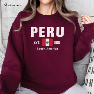 Peut inclure: Un sweat-shirt à col rond bordeaux avec le texte blanc PERU, le texte EST. 1821, un petit graphique du drapeau péruvien et le texte South America. Le mot Maroon est imprimé dans le coin supérieur gauche de l'image en police cursive.
