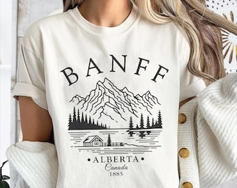 May include: Ivory-colored t-shirt with a black graphic of mountains, trees, and a lake. The word "BANFF" is above the graphic, with "ALBERTA Canada 1885" below. The shirt has rolled-up sleeves.