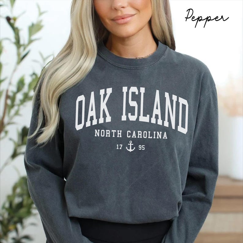 May include: Dark grey sweatshirt featuring the text "OAK ISLAND NORTH CAROLINA 17 95" in white block letters. The sweatshirt has a crew neck and long sleeves. The word "Pepper" is written in cursive in the upper right corner.