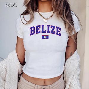 May include: A white short-sleeved t-shirt featuring the word BELIZE in large blue capital letters arched over a small rectangular Belizean flag graphic. The shirt has a crew neckline and is worn with a gold-toned chain necklace and beige trousers.