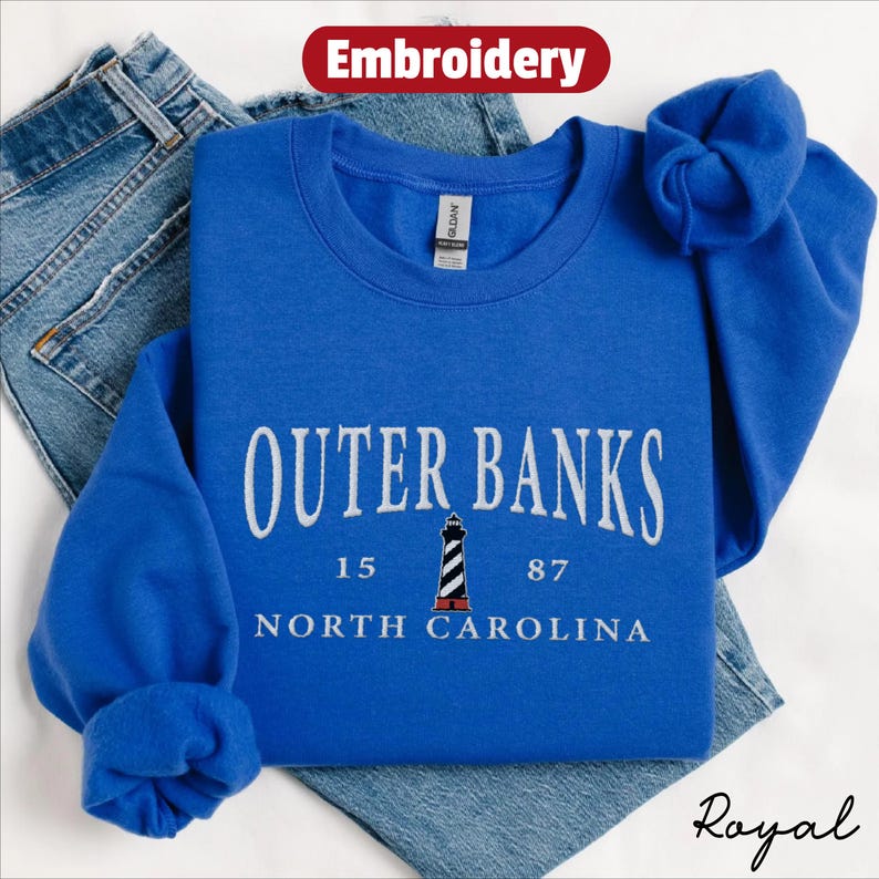 Pode incluir: Um moletom azul royal com as palavras "Outer Banks" em letras brancas arqueadas, juntamente com um gr&aacute;fico de farol e o texto "North Carolina". O moletom est&aacute; dobrado sobre um par de jeans azuis. A palavra "Embroidery" est&aacute; em uma caixa vermelha.