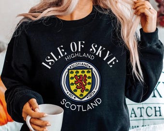 Może przedstawiać: Czarna bluza z długim rękawem z białym napisem ISLE OF SKYE HIGHLAND SCOTLAND. Okrągły emblemat pośrodku przedstawia żółtą tarczę z czerwonym lwem, otoczoną napisem SCOTLAND EST 1873 w niebiesko-białej obwódce.