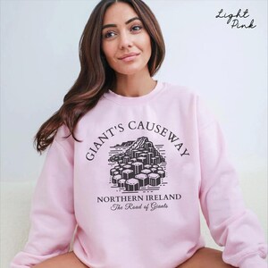 May include: A light pink sweatshirt with a black graphic of the Giant's Causeway in Northern Ireland. The text "Giant's Causeway" is in a black circle around the graphic. The text "Northern Ireland" and "The Road of Giants" is below the graphic.