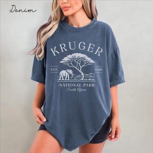 Puede incluir: Camiseta azul denim con un gr&aacute;fico de un elefante, un &aacute;rbol y el texto "KRUGER NATIONAL PARK - South Africa". La camiseta tambi&eacute;n tiene el texto "EST. 1926".