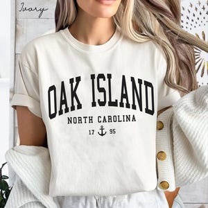 May include: Cream-coloured t-shirt featuring the text "OAK ISLAND" in bold black lettering. Beneath, it reads "NORTH CAROLINA" with the numbers "17 95" and an anchor symbol. The shirt has rolled-up sleeves.