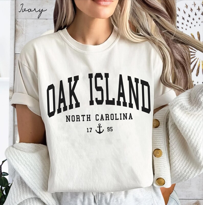May include: Cream-coloured t-shirt featuring the text "OAK ISLAND" in bold black lettering. Beneath, it reads "NORTH CAROLINA" with the numbers "17 95" and an anchor symbol. The shirt has rolled-up sleeves.