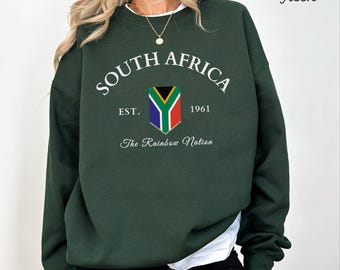 Può includere: Una felpa girocollo verde bosco con testo bianco che recita SOUTH AFRICA, EST. 1961 e The Rainbow Nation. Un grafico a scudo che mostra i colori della bandiera sudafricana è centrato tra il testo. Il tessuto è di un colore verde scuro unito.