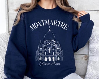 Może przedstawiać: Granatowa bluza z długim rękawem z białą grafiką liniową przedstawiającą bazylikę Sacré-Cœur na Montmartre. Nad budynkiem znajduje się łukowaty napis "MONTMARTRE", a pod ilustracją widnieje napis "France, Paris" wykonany pismem odręcznym.