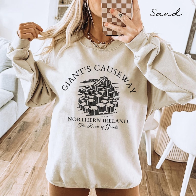 May include: A beige sweatshirt with a black graphic print of the Giant's Causeway in Northern Ireland. The text "Giant's Causeway" is printed in a black circular arc above the graphic. The text "Northern Ireland" is printed below the graphic, and the text "The Road of Giants" is printed below that.