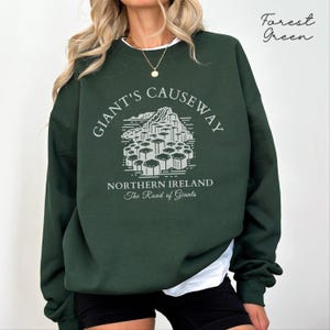 May include: Forest green sweatshirt with a white graphic print of the Giant's Causeway in Northern Ireland. The text "Giant's Causeway" is in a circular design around the graphic. The text "Northern Ireland" and "The Road of Giants" is below the graphic.