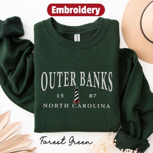 Pode incluir: Camisola verde floresta com o texto branco "OUTER BANKS", um gr&aacute;fico de farol e "NORTH CAROLINA". A palavra "Embroidery" est&aacute; num oval vermelho. As palavras "Forest Green" est&atilde;o na parte inferior.