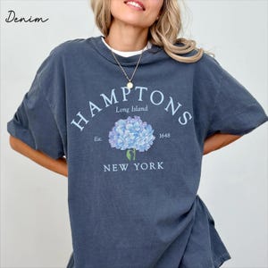 Pode incluir: Uma t-shirt de manga curta azul-escura com um gráfico de hortênsia azul-clara ao centro. O texto diz HAMPTONS, Long Island, Est. 1648 e NEW YORK numa fonte azul-clara. A palavra Denim está impressa no canto superior esquerdo.