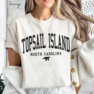 May include: Ivory-colored t-shirt with the text "TOPSAIL ISLAND NORTH CAROLINA" in bold black letters. A black sea turtle graphic is below the text. The shirt has short sleeves and a relaxed fit.
