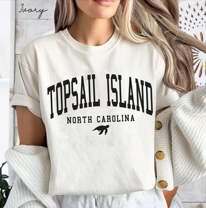 May include: Ivory-colored t-shirt with the text "TOPSAIL ISLAND NORTH CAROLINA" in bold black letters. A black sea turtle graphic is below the text. The shirt has short sleeves and a relaxed fit.