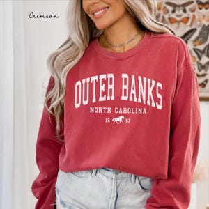May include: Crimson-colored sweatshirt with the text "Outer Banks North Carolina" in white block letters. Below the text is a horse graphic and the numbers "15 87". The word "Crimson" is written in cursive in the upper left corner.