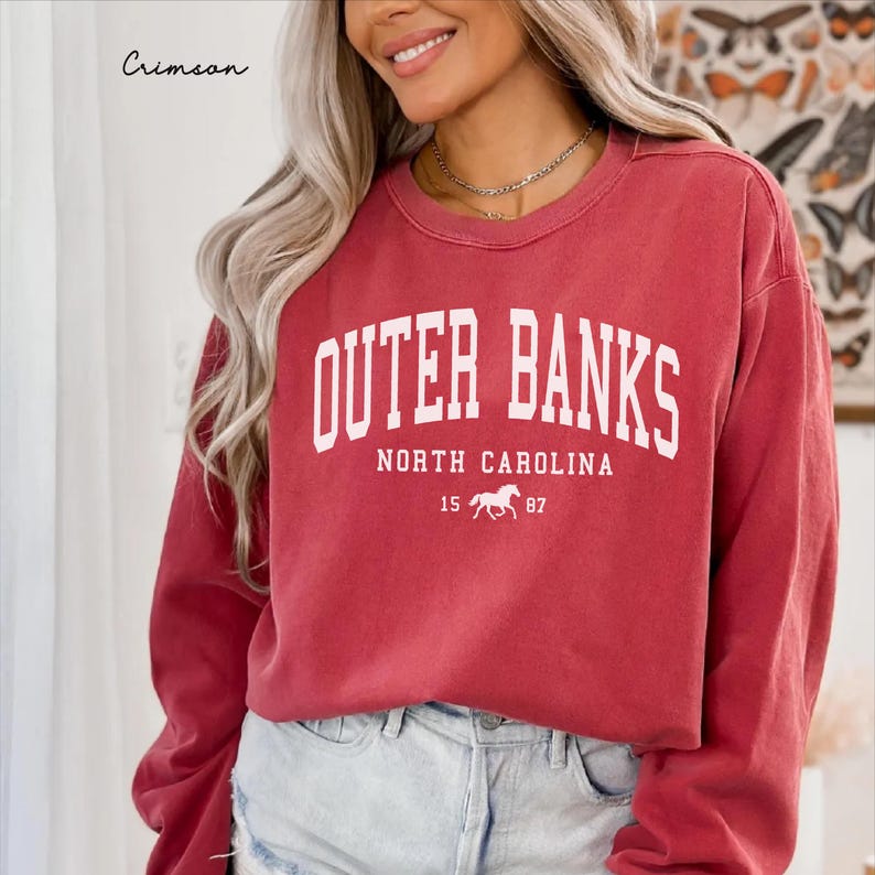 May include: Crimson-colored sweatshirt with the text "Outer Banks North Carolina" in white block letters. Below the text is a horse graphic and the numbers "15 87". The word "Crimson" is written in cursive in the upper left corner.