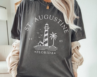Może przedstawiać: Ciemnoszara koszulka z białym nadrukiem latarni morskiej, palmy i napisem "ST. AUGUSTINE FLORIDA". Koszulka ma wygląd vintage.