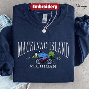 May include: Navy blue sweatshirt with embroidered "Mackinac Island" text and a bicycle graphic. The design includes the years "1780" and the state "Michigan". A silver necklace with a pendant is visible.