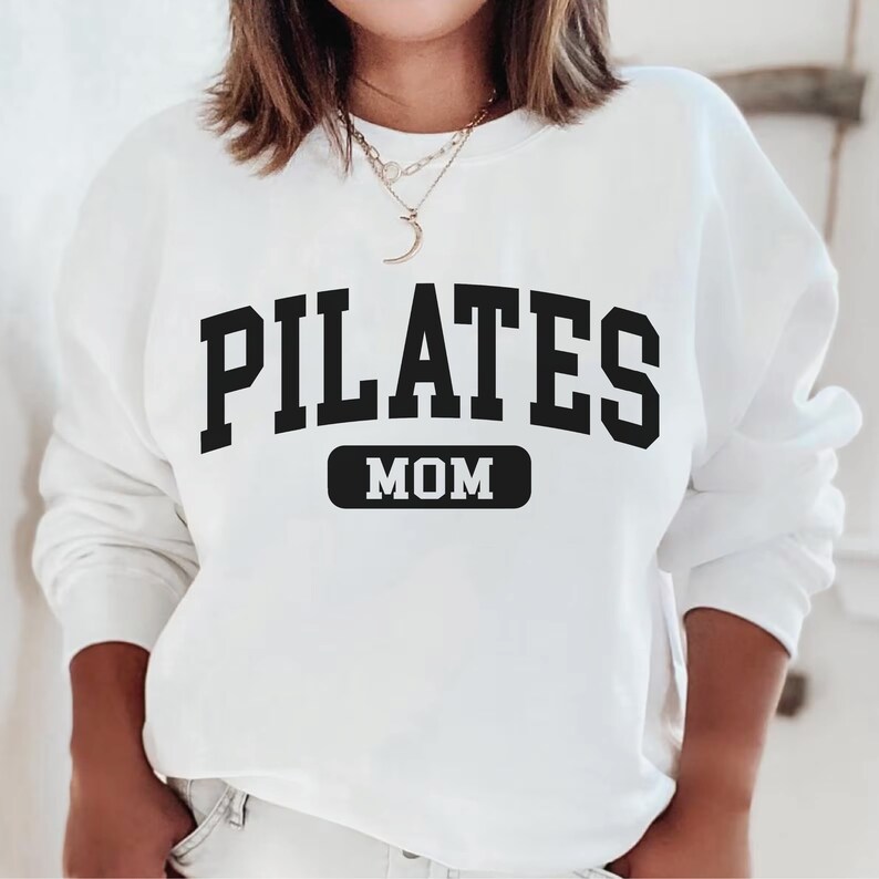 Pu&ograve; includere: Felpa bianca con testo nero che recita "PILATES MOM" in un font da college.