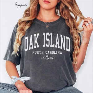 May include: Dark grey t-shirt featuring the white text "OAK ISLAND NORTH CAROLINA". Below the text are the numbers "17 95" with an anchor symbol. The word "Pepper" is in the upper left corner of the image.