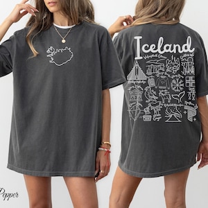 May include: Dark gray t-shirt with a white outline of Iceland on the front. The back of the shirt features the word "Iceland" above a collection of white illustrations of Icelandic landmarks and symbols. The shirt is a relaxed fit.
