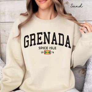 Może przedstawiać: Beżowa bluza z napisem "GRENADA" pogrubionymi czarnymi literami. Poniżej napis "SPICE ISLE" z flagą Grenady i liczbami "19 74". Bluza ma okrągły dekolt.