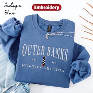 Pode incluir: Camisola azul &iacute;ndigo com gola redonda com o texto "Outer Banks" em branco, com um gr&aacute;fico de farol. A camisola tamb&eacute;m inclui o texto "15 87 North Carolina". Um colar de prata est&aacute; drapeado sobre a camisola.