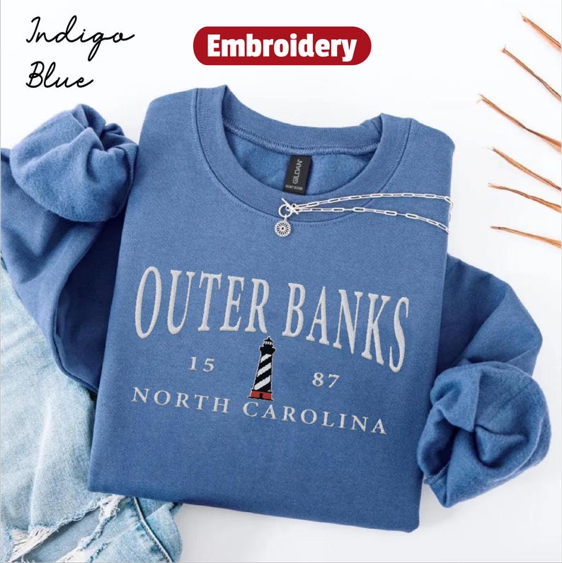 Pode incluir: Camisola azul &iacute;ndigo com gola redonda com o texto "Outer Banks" em branco, com um gr&aacute;fico de farol. A camisola tamb&eacute;m inclui o texto "15 87 North Carolina". Um colar de prata est&aacute; drapeado sobre a camisola.