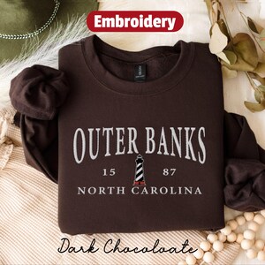 Pode incluir: Camisola castanho-escura com texto bordado branco "Outer Banks, Carolina do Norte". Um gr&aacute;fico de farol est&aacute; centrado abaixo do texto. A palavra "Embroidery" est&aacute; em uma faixa vermelha no topo. As palavras "Dark Chocolate" est&atilde;o na parte inferior.