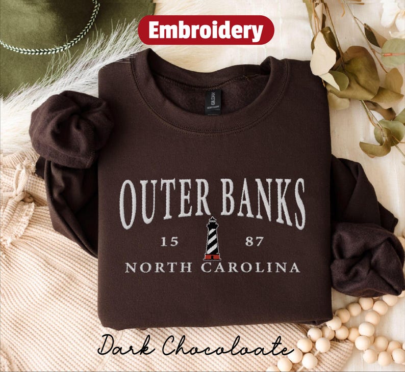 Pode incluir: Camisola castanho-escura com texto bordado branco "Outer Banks, Carolina do Norte". Um gr&aacute;fico de farol est&aacute; centrado abaixo do texto. A palavra "Embroidery" est&aacute; em uma faixa vermelha no topo. As palavras "Dark Chocolate" est&atilde;o na parte inferior.