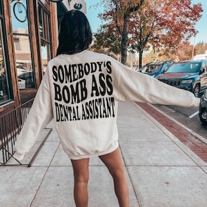 Op de afbeelding: Een wit sweatshirt met de tekst "Somebody's Bomb Ass Dental Assistant" in het zwart gedrukt op de achterkant.