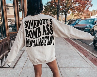 May include: A white sweatshirt with the text "Somebody's Bomb Ass Dental Assistant" printed in black on the back.