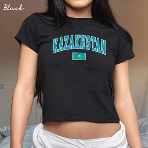 May include: A black short-sleeved t-shirt featuring the word KAZAKHSTAN in a bold, arched, light blue collegiate-style font. Below the text is a small, rectangular graphic of the national flag of Kazakhstan, which is light blue with a yellow sun and eagle.