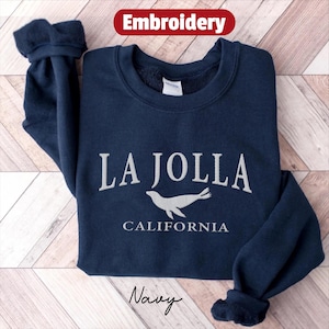 May include: Navy blue sweatshirt with the embroidered text "LA JOLLA CALIFORNIA" and a seal graphic. The word "Embroidery" is in a red banner at the top. The word "Navy" is at the bottom.