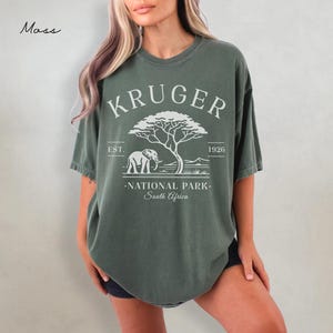 Puede incluir: Camiseta verde musgo con un gr&aacute;fico blanco de un elefante, un &aacute;rbol y el texto "KRUGER NATIONAL PARK - South Africa". La camiseta tiene un aspecto vintage y descolorido.