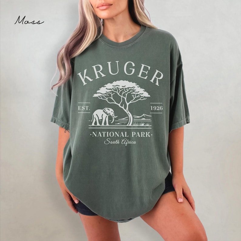 Puede incluir: Camiseta verde musgo con un gr&aacute;fico blanco de un elefante, un &aacute;rbol y el texto "KRUGER NATIONAL PARK - South Africa". La camiseta tiene un aspecto vintage y descolorido.