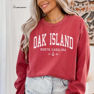 May include: Crimson red sweatshirt with the text "Oak Island North Carolina" in white block letters. Below the text is the number "17" and "95" with an anchor symbol. The word "Crimson" is written in cursive in the upper left corner.