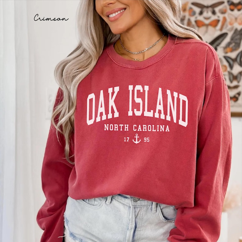 May include: Crimson red sweatshirt with the text "Oak Island North Carolina" in white block letters. Below the text is the number "17" and "95" with an anchor symbol. The word "Crimson" is written in cursive in the upper left corner.