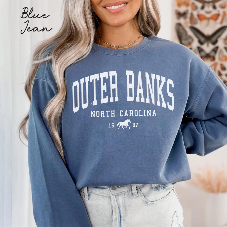 May include: Blue jean-colored sweatshirt with the text "Outer Banks North Carolina" in white block letters. Below the text is a white graphic of a horse and the numbers "15 87". The sweatshirt has long sleeves.