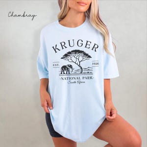 Puede incluir: Camiseta azul claro con un gr&aacute;fico negro de un elefante, un &aacute;rbol y las palabras "KRUGER NATIONAL PARK South Africa". La camiseta tambi&eacute;n tiene el texto "EST. 1926" encima y debajo del gr&aacute;fico. La palabra "Chambray" est&aacute; escrita en cursiva en la esquina superior izquierda.